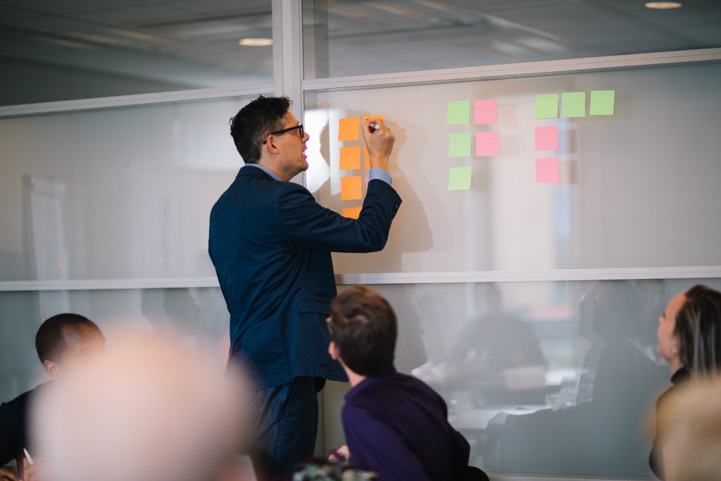 Mann schreibt etwas auf ein Post-It an einer Tafel